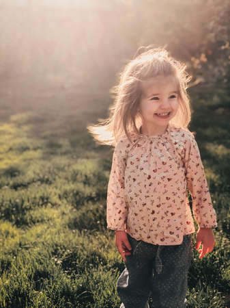 Close up photo of smiling cute little girl of 3-4 years old with blond fluffy hair stands outdoor in...
