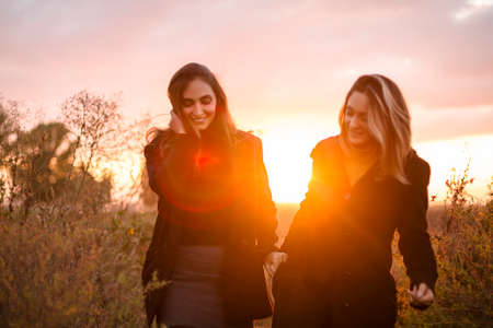 Two happy women girlfriends holding hands walking through autumn field at sunset and smiling,...