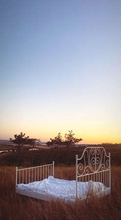 Landscape photo shoot in nature, wrought iron unmade empty bed with white linen standing in summer evening field with dry yellow tall grass at sunset. Healthy night sleep and relaxation conceptの写真素材