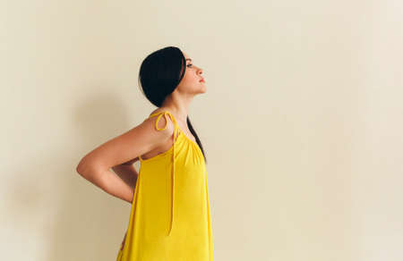 Calm young woman in summer yellow dress isolated over beige background, copy space for text, Tired female suffering from backache or back pain, massaging lower back by herself. side viewの写真素材