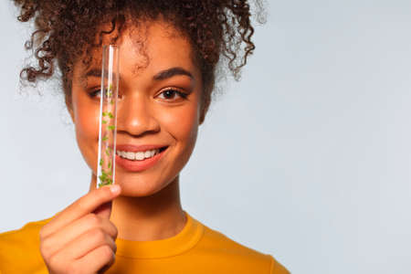 Smiling african american woman holding glass test tube with microgreens in front of face while posing isolated over gray background, copy space for text. Superfood and nutrition conceptsの写真素材