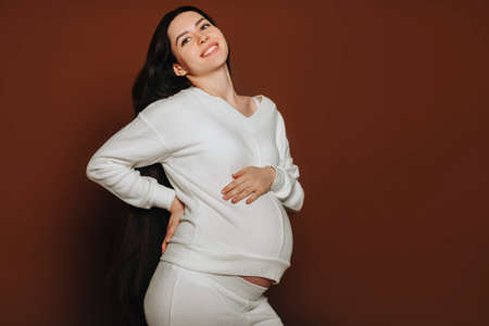 Young happy pregnant woman with long brown hair touching belly, talking with unborn baby while...