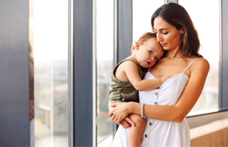Being mom. Young beautiful woman mother holding cute baby boy while standing by window at home....