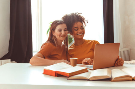Two happy girlfriends preparing for exams, sitting together in front of laptop next to open textbooks against backdrop of living room interior, cheerful classmates students studying teamwise indoorの写真素材