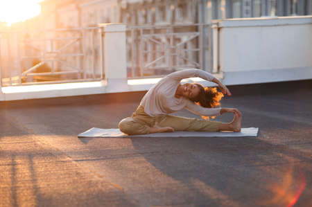 Young sporty happy mixed-race woman in sportswear doing stretching exercises while sitting on yoga mat on house roof in early morning, joyful fit female exercising at sunriseの写真素材