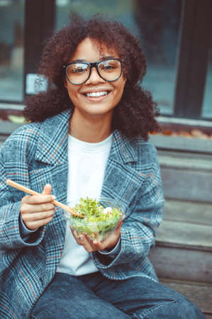 Happy african american woman office worker eating salad and smiling at camera while sitting on bench in park outdoors, selective focus. Positive black girl having lunch outside during break at workの写真素材