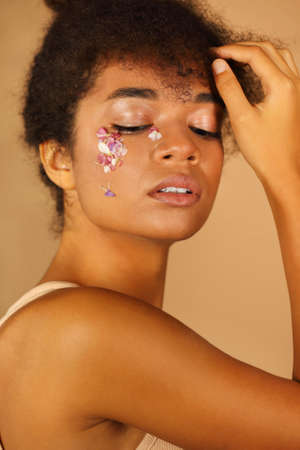 Portrait of beautiful African girl with curly hair shows creative make-up with flower petals while hand gently touches her face isolated over beige background. Natural eco cosmetics and beauty conceptの写真素材