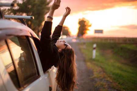 Young carefree woman traveler stretching out of car window while driving through countryside on beautiful summer evening at sunset, happy female travelling by car in nature. Travel and road tripの写真素材