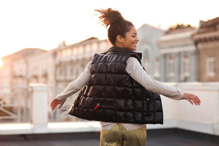 Young happy carefree african american woman dancing having fun at rooftop in morning sunlight with cityscape on background, happy mixed-race female in casual clothes moving to music on roofの写真素材