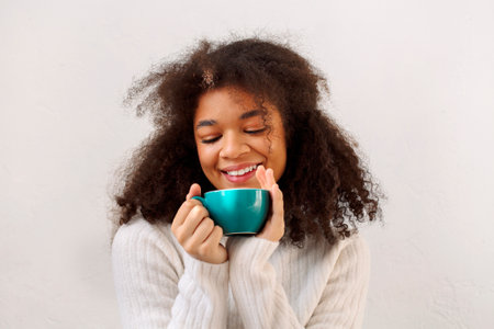 Happy pleased delighted curly african american girl with closed eyes enjoying cup of tea and relaxing in cold winter season, wearing warm knitted white sweater, isolated on white studio background.の写真素材