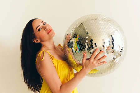 Portrait of young brunette woman with long hairstyle and bright makeup holding in hands big shiny and glossy disco ball which reflects everything around, standing against white studio wall backgroundの写真素材