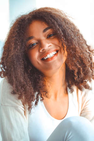 Pleased relaxed African American ethnicity girl in homewear sitting on windowsill against backdrop...