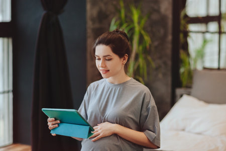 Young smiling pregnant woman holding digital table and making video call while sitting on bed at...