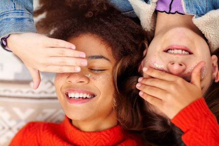 View from above of two happy multiracial teen girls covering eyes and smiling while lying on blanket...