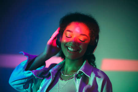 Young happy african american woman in black headphones listening to music, enjoying high quality sound while standing under purple neon glowing light in studio and smiling happily. musical equipmentの写真素材