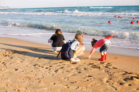 Photo from behind of joyful family standing on seashore looking at surf and waves, mom and two...