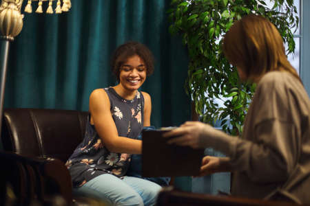 African-American young woman smiling while sitting in psychologist office. Female psychotherapist offering client to take test to determine cause of anxiety and find helping solution. selective focusの写真素材
