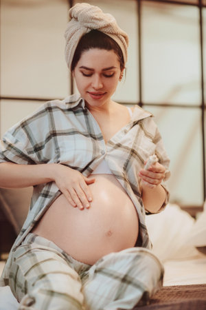 Cute young pregnant woman wearing pajama and wrapped towel on head applying body lotion on her belly in shape of heart and smiling at camera, enjoying beauty routine at home. Skincare during pregnancyの写真素材