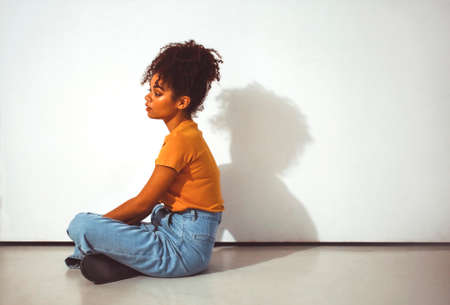 Young calm pensive african american girl with high puff hairstyle sitting in lotus pose on floor against white wall with shadow, thoughtful black woman in casual clothes thinking about life. side viewの写真素材