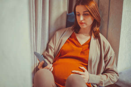 Pregnant woman holding smartphone using pregnancy tracker app on mobile phone while sitting at table...