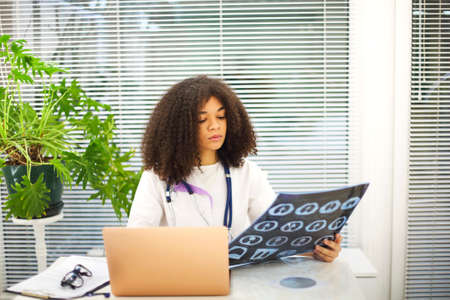 Portrait of ethnic female doctor analyzing X-ray or MRI scan while working in hospital, selective...