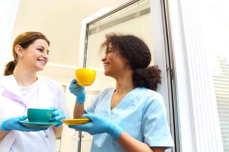 Two happy multi-ethnic female medical workers colleagues drinking coffee or tea and talking while standing near window in modern clinic or hospital, taking a break from workの写真素材