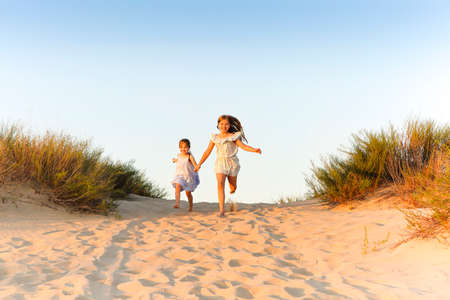 Two happy cheerful little sisters running hand in hand on sand on sunny evening beach, funny girls laughing and smiling spending time with family on seashore. travel and summer vacation conceptの写真素材