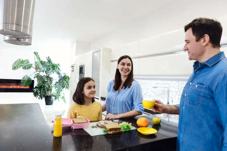 Happy smiling young family enjoying healthy breakfast together with there little daughter at modern white kitchen cooking lunch box for school. care and love conceptの写真素材