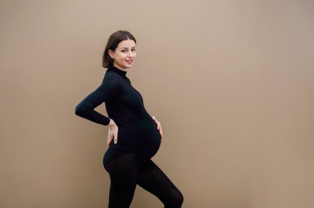 Young pregnant woman standing against beige background looking to the camera touching her tummy wearing black clothesの写真素材