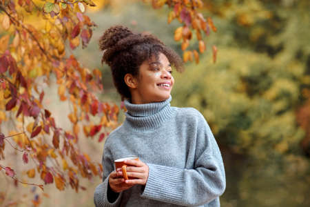 Young happy smiling mixed-race woman with coffee cup in autumn nature, pleased african american female with curly hair in knitted sweater looking away while enjoying morning in beautiful fall gardenの写真素材