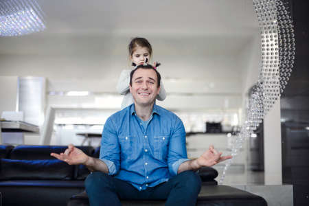 Young brunette man with his cute little daughter at home. Cheerful father playing with daughter in modern apartment together and getting funny creative hairdressの写真素材