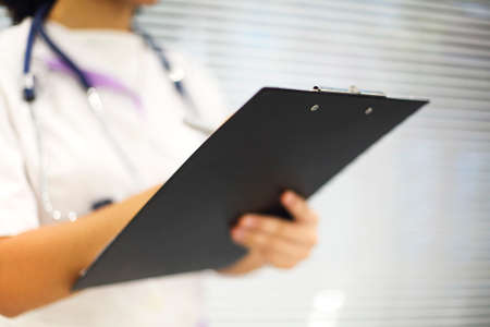 Ethnic female medic in mask and uniform filling medical report on clipboard in modern room in clinic. healthcare conceptの写真素材