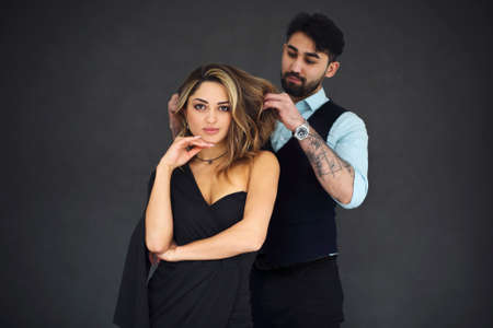 Stylish young ethnic couple embracing against dark gray wall background looking to camera...