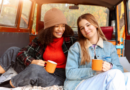 Two happy multiracial girlfriends on car trunk in fall forest, embracing and drinking hot tea during adventure trip in autumn nature, smiling female friends enjoying life while traveling togetherの写真素材