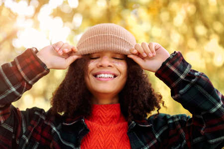 Young happy overjoyed african american woman with curly hair adjusting knitted beige hat and laughing while standing in autumn forest, carefree mixed-race female enjoying fall nature in parkの写真素材