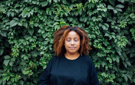 Young positive african woman smiling outside with a ivy plant in the background at street of city wearing on black blouse and blue jeans. Calm and nature in the city conceptの写真素材