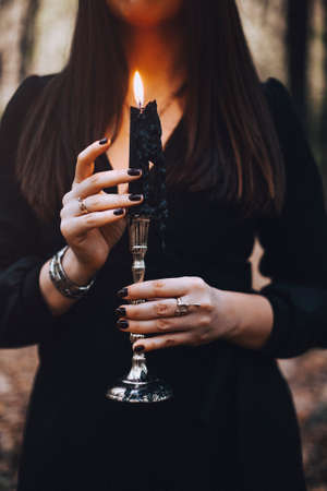 Cropped shot of mysterious woman in black dress holding candle on candlestick in her hands...