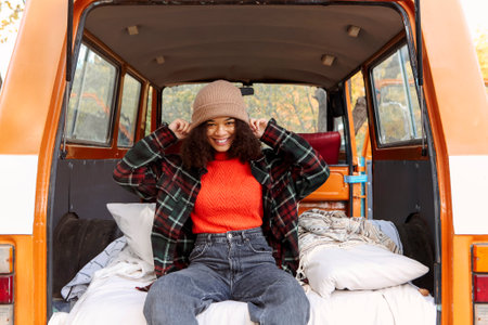 Happy joyful african american young woman traveler sitting in minivan in forest, smiling mixed-race female relaxing inside of orange old vintage van and enjoying adventure trip in autumn natureの写真素材