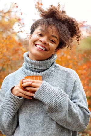 Young happy smiling mixed-race woman with coffee cup in autumn nature, pleased african american female with curly hair in knitted sweater looking attye camera while enjoying morning in beautiful fall gardenの写真素材