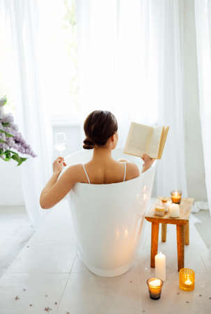 Back view of the young brunette woman reading the book and drinking white wine in bath with foam and candlesの写真素材