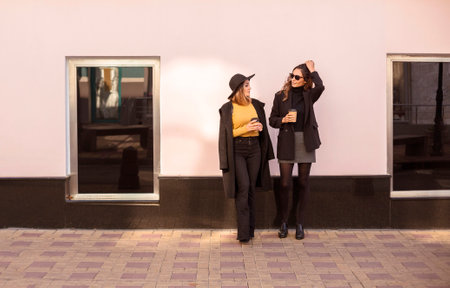 Two stylish women female friends in trendy clothes enjoying nice talk and drinking take away coffee...