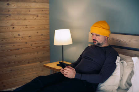 Portrait of mature handsome businessman sitting in yellow hat in hotel in rustic style with mobile...