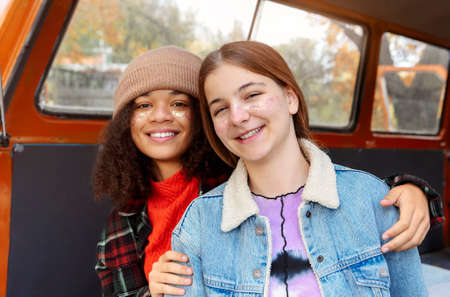 Picnic in autumn nature. Two happy multiracial girlfriends sitting on car trunk in forest, raising arms smiling at camera during adventure road trip on fall season, smiling female friends in minivanの写真素材