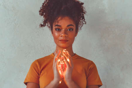Happy african american girl standing with festive led lights around hands and body, raising up one...