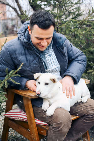 Man and Cute white Dog Sitting on Chairs At Park At Sunny Winter Day. Concept of Friendship Between...
