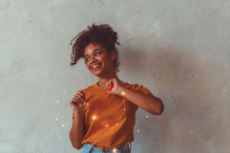 Happy african american girl standing with festive led lights around hands and body, raising up one...