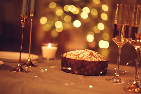 Sliced of freshly baked panettone cake lying on wooden cutting board on table served for festive dinner with clean plates, burning candles, led lights and glasses. rustic styleの写真素材