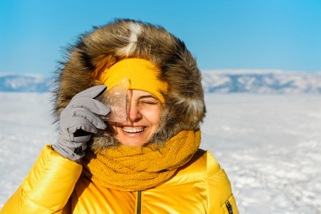 Happy smiling woman wearing winter yellow jacket holding piece of transparent crushed ice cubes over...