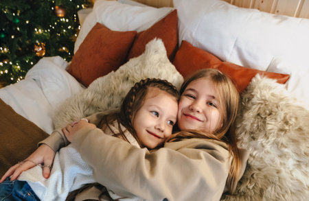 Cozy domestic portrait of two happy smiling little girls sisters hugging embracing while spending leisure time together at home, lying on bed near decorated Christmas tree. family and children conceptの写真素材