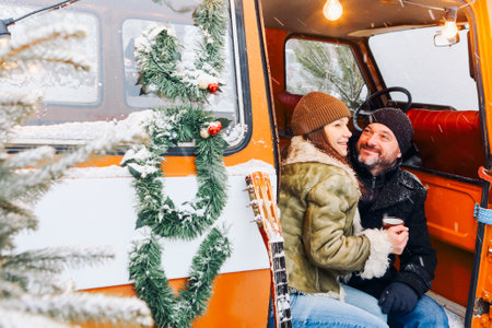 Happy middle-aged romantic couple in love sitting in minivan decorated with coniferous branches, drinking hot tea. Man and woman in warm clothes relaxing in nature during winter holidaysの写真素材